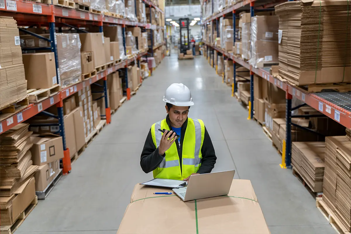 Warehouse worker in safety vest and hard hat using a two-way radio at a desk with a laptop and papers.