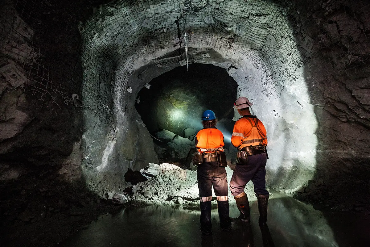 Two workers in safety gear standing in a reinforced mining tunnel illuminated by headlamps, with rubble on the ground.