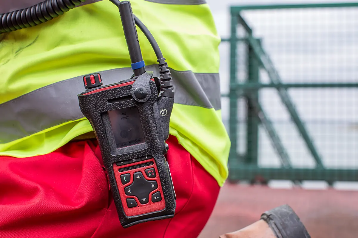 Close-up of a TETRA radio device on a worker’s belt, with high-visibility clothing and an industrial site in the background.