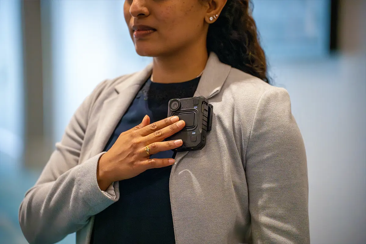 Person in a grey blazer with a body worn camera attached to the chest, hand activating the device.
