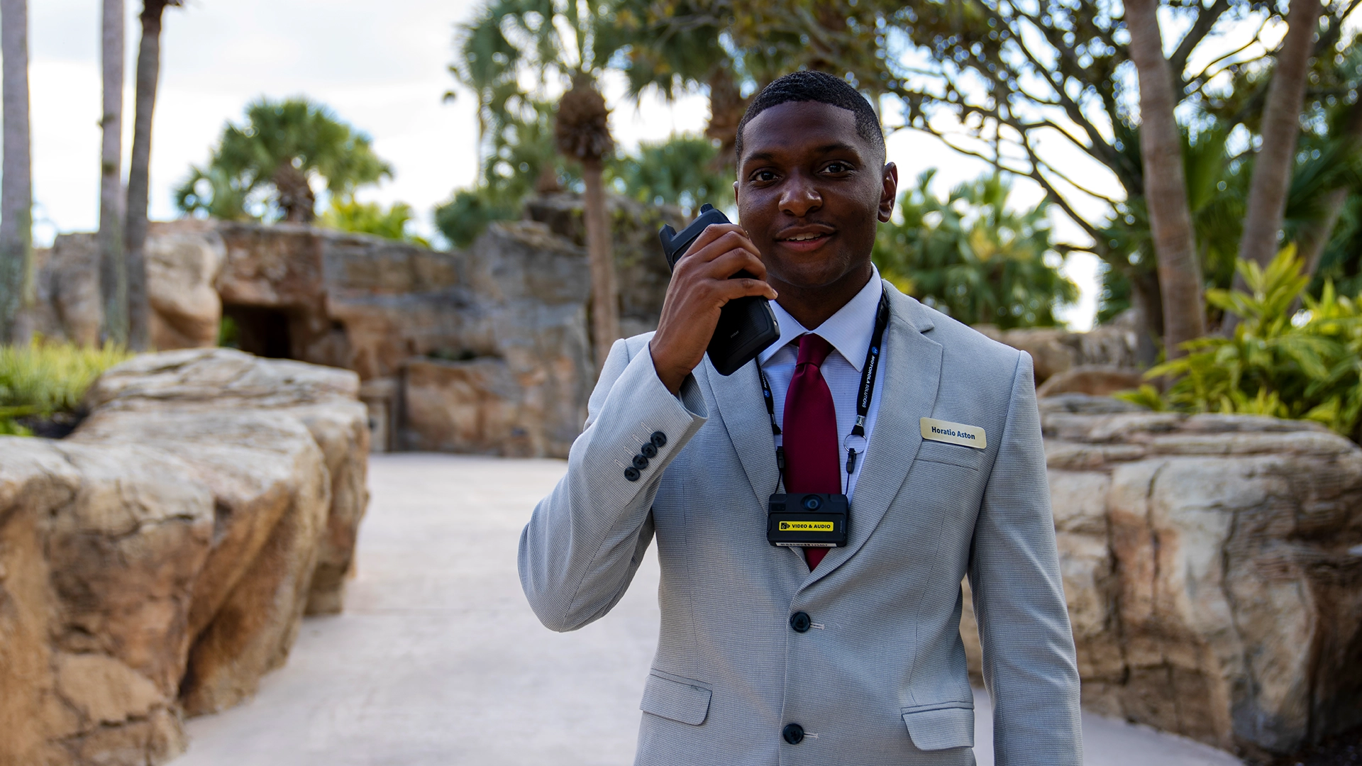 A suited professional using a two-way radio in an outdoor setting with rocks and palm trees, representing Logic Technologies’ business-critical communication solutions.