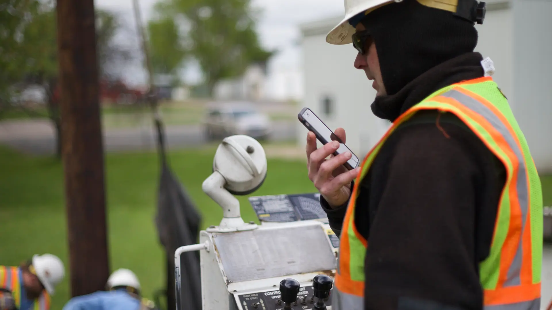 Worker in safety gear using a smartphone near industrial equipment, with other workers visible in the background.