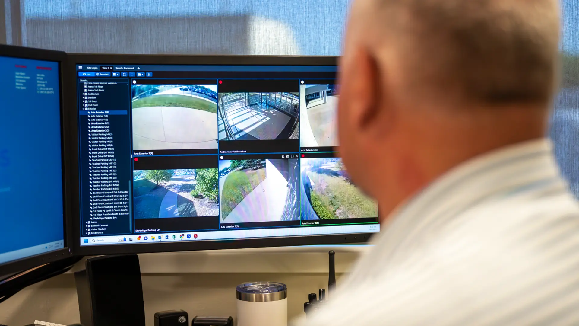 Person viewing a computer monitor displaying multiple security camera feeds of outdoor areas and building entrances.