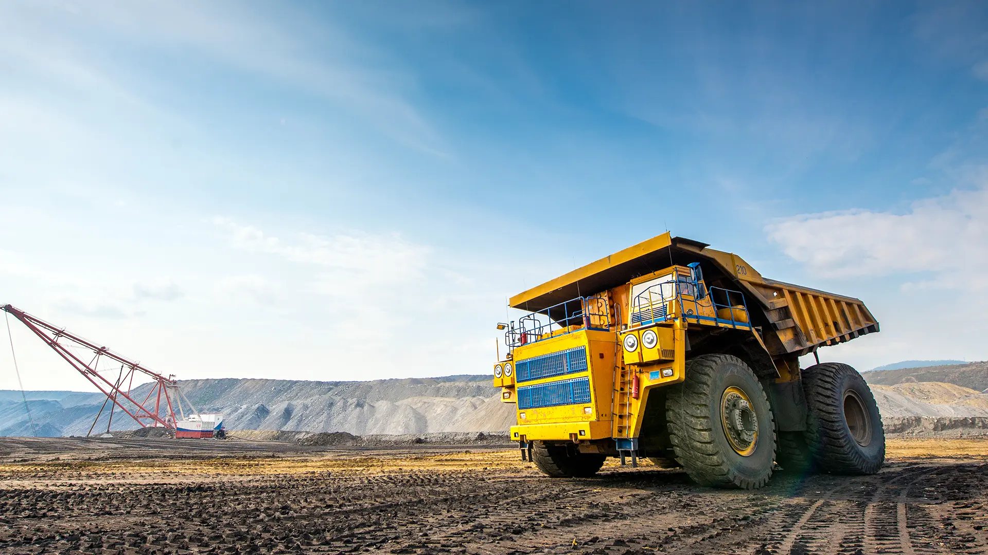 Large mining truck and excavator in an open-pit mine with exposed earth layers and rugged terrain.