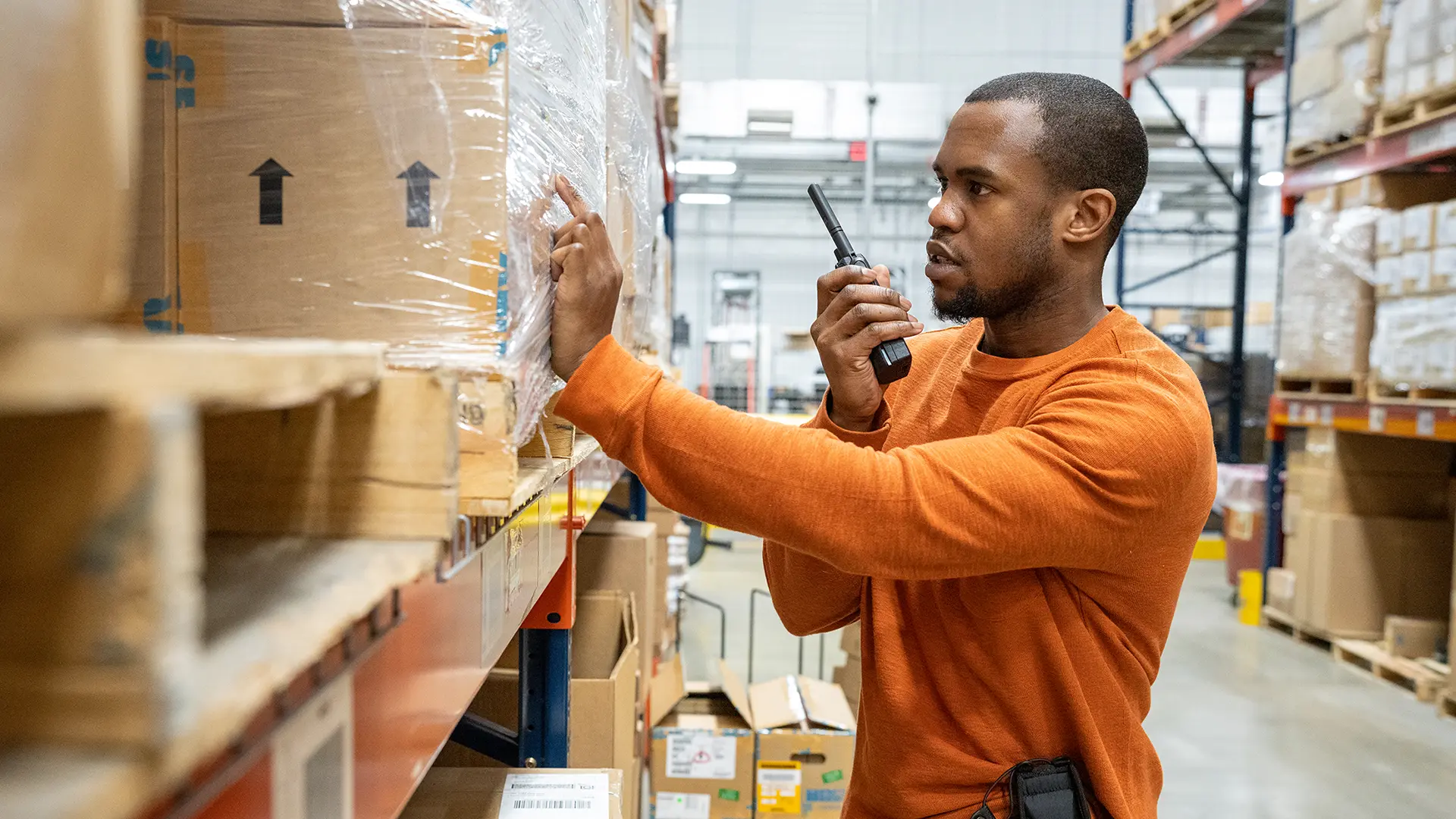 Warehouse worker in orange shirt using a two-way radio beside shelves filled with boxes and equipment.