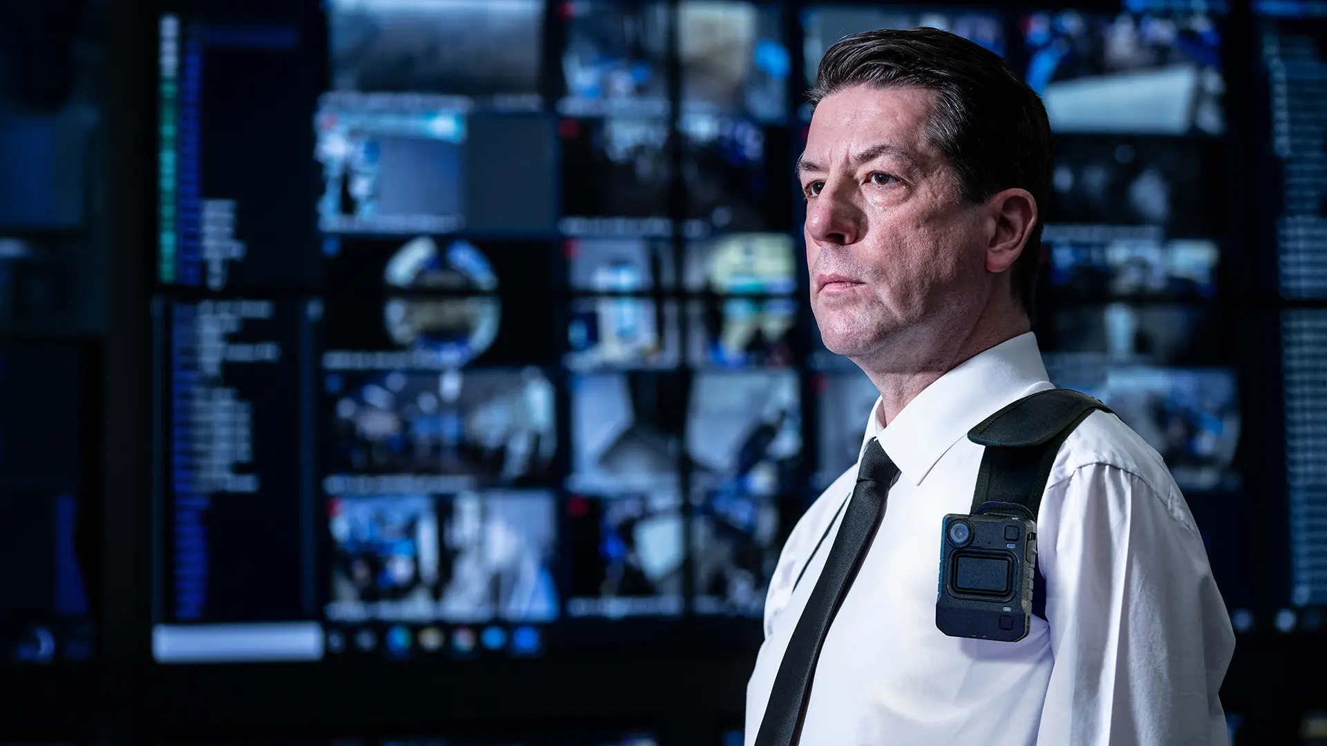 Person wearing a body camera in a security control room with multiple screens displaying video feeds and information.